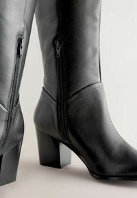 Bottes en cuir noir montantes jusqu'au genou avec un talon carré, dotées de fermetures éclair sur les côtés et d'une texture lisse. Coutures détaillées visibles.