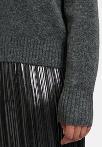 Dunkelgrauer gestrickter Pullover mit gerippten Bündchen, kombiniert mit einem glänzenden schwarzen Faltenrock, der einen kontrastierenden Textur- und Glanzeffekt zeigt.