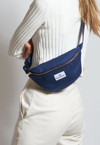 Riñonera de lona azul marino con un zipper dorado y un parche de logo blanco, llevada a través de la cintura. Textura suave y diseño curvado.