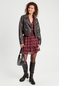 Veste de biker en cuir noir courte sur une robe rouge à motifs, accompagnée de bottes noires jusqu'aux genoux et d'un petit sac à main noir.