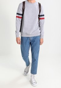 Grauer Sweatshirt mit roten und marineblauen Streifen an den Ärmeln, kombiniert mit hellblauen Jeans und weißen Sneakern, während ein brauner Rucksack getragen wird.