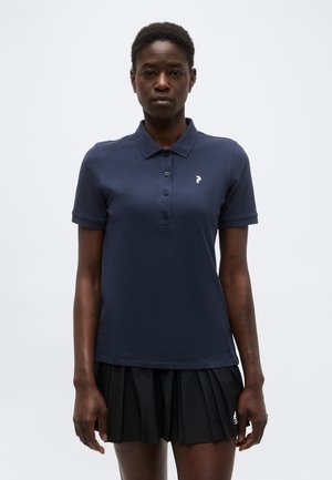 Femme portant un polo bleu marine à manches courtes avec des boutons et une jupe plissée noire sur un fond uni.