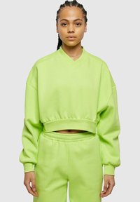 Felpa verde neon corta con scollo a V, maniche ampie e orlo elastico. Abbinata a pantaloni della tuta a vita alta coordonati.