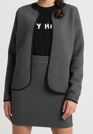 Kobieta ubrana w teksturowaną, ciemnoszarą dzianinową kurtkę i dopasowaną spódnicę, na czarnej koszuli z częściowym białym nadrukiem, stojąca na jednolitym tle.