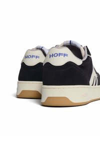 Sneaker con una toma superiore in suede scuro, dettagli in pelle color crema, logo blu "HOFF" e suola in gomma testurizzata con un design semplice.