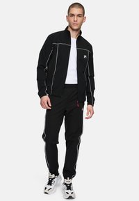 Zwart sportief trainingspak met witte piping, een jacket met rits, broeken met elastische tailleband en logo-detail op de borst. Gedragen met witte sneakers.