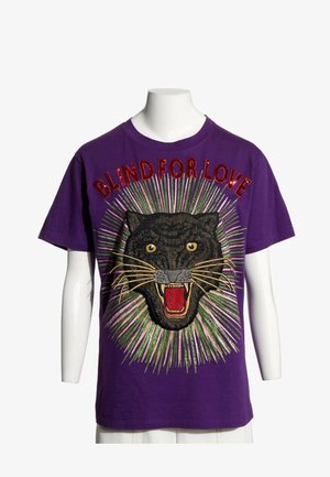 Camiseta morada con un gran pantera negra rugiente bordada y texto rojo que dice "BLIND FOR LOVE" encima.