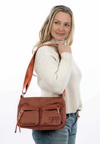 Borsa in tessuto color ruggine con due tasche anteriori con zip e accenti laterali. Presenta una tracolla ampia e regolabile e una superficie texture.