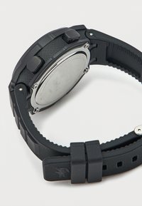 Μαύρο ρολόι από καουτσούκ με υφή στη λωρίδα, ασημένιο μεταλλικό εξάρτημα και δύο κουμπιά. Η πίσω πλευρά έχει γυαλιστερή μεταλλική επιφάνεια με λογότυπο.