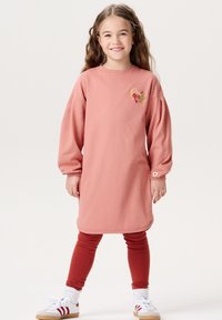 Rosa Sweatkleid mit langen Ärmeln und rundem Ausschnitt. Verziert mit einem bestickten Design auf der Brust. Kombiniert mit roten Leggings und Sneakers.