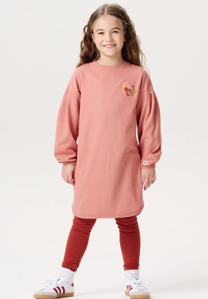 Rosa Sweatkleid mit langen Ärmeln und rundem Ausschnitt. Verziert mit einem bestickten Design auf der Brust. Kombiniert mit roten Leggings und Sneakers.