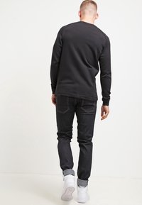 Svart långärmad tröja med rund halsringning, i kombination med mörka jeans och vita sneakers, visad bakifrån. Slät textur, enkel design.