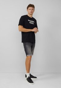Zwarte katoenen t-shirt met de tekst "EVOLVING ROOTS", gecombineerd met denim shorts met opgerolde zoom in het grijs en zwarte sneakers met witte zolen.