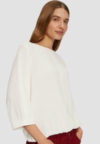 Haut texturé blanc avec un col rond, des manches trois-quarts et un ourlet froncé, associé à un pantalon en velours côtelé rouge.