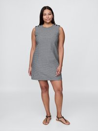Jeune femme debout, portant une robe sans manches à carreaux noir et blanc et des sandales plates noires, devant un fond clair et uni.