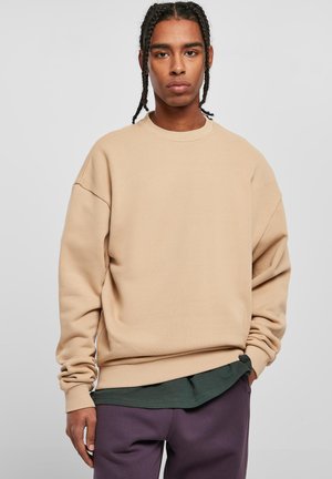 Beige hettegensere laget av mykt stoff, med rund hals, senkede skuldre og ribbede mansjetter. Brukt over en mørkegrønn skjorte og lilla bukser.