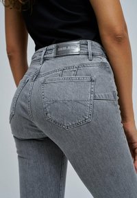 Primer plano de una persona que lleva jeans grises de talle alto con bolsillos traseros y una etiqueta de cuero de la marca "Salsa Jeans" en la cinturilla.