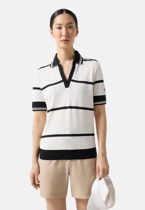 Kvinde iført en hvid kortærmet polo med sorte striber, beige shorts, holder en hvid solskærm i sin højre hånd.