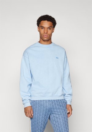 Sweat-shirt bleu clair avec un col rond et des poignets côtelés, présentant un petit logo sur la poitrine. Associé à un pantalon bleu à motifs.