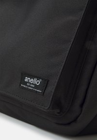 Svart tygväska med en texturerad yta. Har en anello®-etikett som visar grundat 2005, och en hållbar dragkedjestängning.