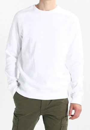 Person trägt ein schlichtes weißes Sweatshirt mit langen Ärmeln und olivgrüne Cargohosen, steht mit einer Hand in der Tasche vor einem weißen Hintergrund.