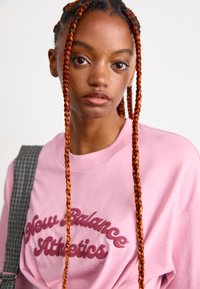 Jeune femme avec de longues tresses rougeâtres portant un sweat-shirt rose New Balance Athletics et une sangle grise à carreaux sur son épaule.