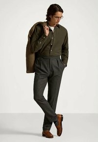 Homem com uma camisa verde oliva abotoada, calças pregueadas cinza-escuras, sapatos de couro castanho, segurando um casaco castanho sobre o ombro.