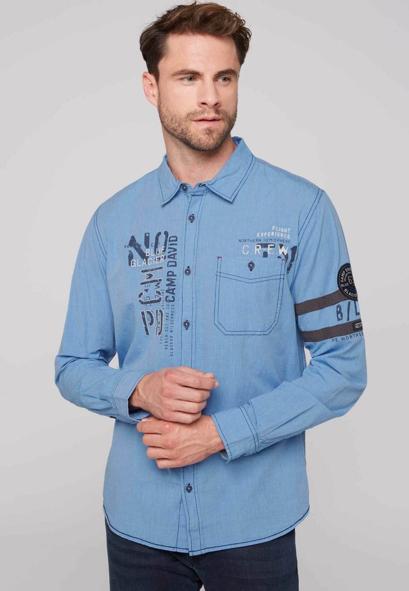 Camisa de manga larga azul claro con cierre de botones, que presenta texto impreso y un parche en la manga izquierda, además de un bolsillo en el pecho.