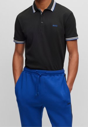 Schwarzes Polohemd mit geripptem Kragen, der blaue und graue Akzente aufweist, kombiniert mit blauen Jogginghosen. Das Hemd zeigt ein dezentes Logo auf der Brust.