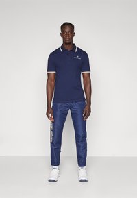 Marineblå polo skjorte med hvite krage striper og logo; kombinert med matchende marineblå bukser med gule detaljer og avsmalnede mansjetter. Hvide sneakers.