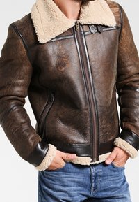 Veste en cuir marron avec un col et des poignets en peau lainée beige. Dispose d'une fermeture éclair frontale et de poches latérales. Surface texturée avec une apparence robuste.
