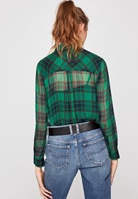 Blusa transparente de cuadros verdes con mangas largas, que presenta un cuello clásico y un detalle de yugo en la parte posterior, combinada con unos jeans de tiro alto en azul y un cinturón negro.