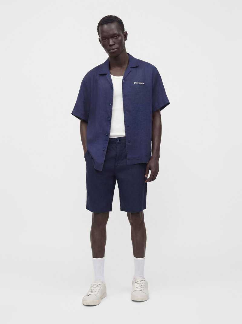 Man wearing navy blue short-sleeve button-up shirt and matching shorts, white tank top, white socks, and white sneakers, standing with hands in pockets.