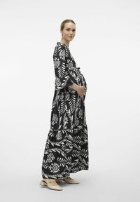 Robe de maternité à motifs noir et blanc avec de longues manches évasées et un ourlet volanté, associée à des chaussures à talons ouverts de couleur claire.