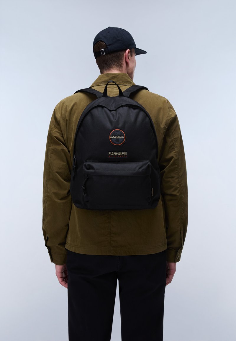 Mann trägt schwarze Mütze, olivgrüne Jacke, schwarze Hose und schwarzen Rucksack mit Vordertasche und rundem Logo vor einfarbigem Hintergrund.