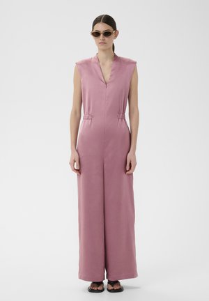 Femme aux cheveux foncés en queue de cheval basse, portant des lunettes de soleil marron et une combinaison rose sans manches à jambes larges avec des sandales noires, debout.