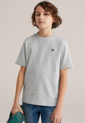 Junge Person mit welligem, kurzem braunem Haar trägt ein hellgraues T-Shirt mit kleinem Vogel-Logo und hält eine grüne Jacke in der linken Hand.