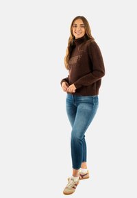 Maglione marrone lavorato a maglia con un alto collo, caratterizzato da un logo. Abbinato a jeans blu aderenti e sneaker beige con accenti viola.