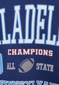 T-shirt azul marinho com texto branco e azul claro, "PHILADELPHIA CHAMPIONS", acompanhado de um gráfico de futebol cinza e detalhes vermelhos.