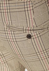 Béžové kárované kalhoty s texturovanou látkou s hnědými a červenými akcenty, přední kapsou a hnědým knoflíkovým detailem.
