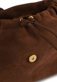 Braune Wildleder-Tasche mit Kordelzug, verziert mit goldfarbenen Metallakzenten und einer glatten Textur, gesichert durch eine dünne schwarze Schnur und einen Druckknopfverschluss.