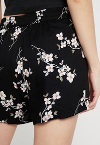Svarta shorts med blommönster som har vita blommor och gröna blad. Elastisk midja, slät textur och lätt rundad nederkant.