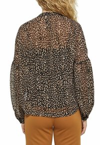 Blusa nera trasparente con stampa animale marrone e beige, caratterizzata da maniche a sbuffo e dettaglio a forma di occhiello nella parte posteriore. Indossata sopra pantaloni arancioni.