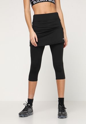Frau trägt schwarze kurze Leggings mit angebrachtem Rock, schwarzen Sport-BH, schwarzen Socken und schwarzen Sportschuhen, steht vor weißem Hintergrund.