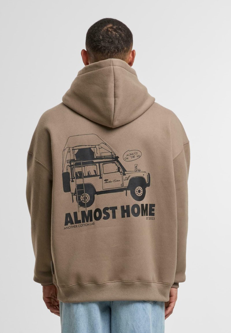 Pull-over en peluche marron avec un grand graphique noir d'un véhicule équipé d'une tente de toit et le texte « PRESQUE CHEZ SOI » au dos.