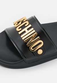 Sandale slide noire à finition brillante, avec une bande accentuée d’un logo doré et une semelle texturée pour l’adhérence. Comprend un détail rond en métal doré.
