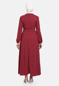 Lange bordeaux jurk met lange mouwen, een tailleband en een vloeiende rok. Gladde stof met een knoopsluiting aan de achterkant.