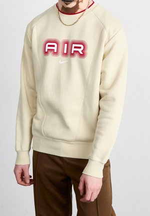 Mężczyzna w kremowej bluzie z czerwonym napisem "AIR" i białym logo Nike, w połączeniu z brązowymi spodniami i złotym łańcuszkiem na szyi.