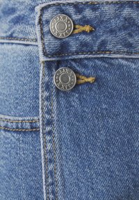 Blue denim jeans featuring two silver metal buttons at the waist, yellow stitching, and a close-up of the textured fabric.