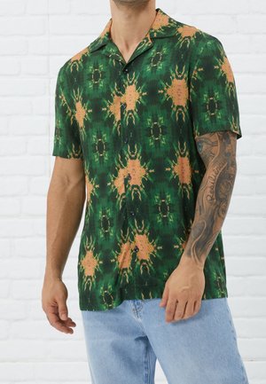 Mand iført grøn og orange mønstret kortærmet skjorte med knapper, lyseblå jeans og synligt tatoveringsærme på højre arm.
